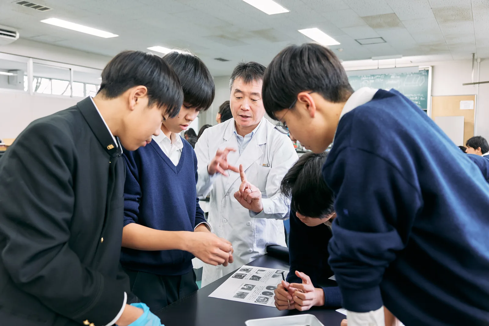 獨協埼玉中学高等学校の理科の授業。白衣の教員を囲んで生徒が手元の資料をのぞき込んでいる