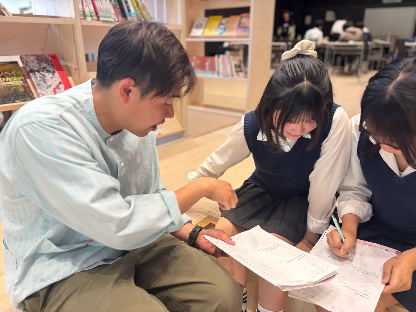 自由学園中等部・高等部の「探求」の時間。本棚のある空間で、大人1人と生徒2人が床に座り、プリントを見ながら話している