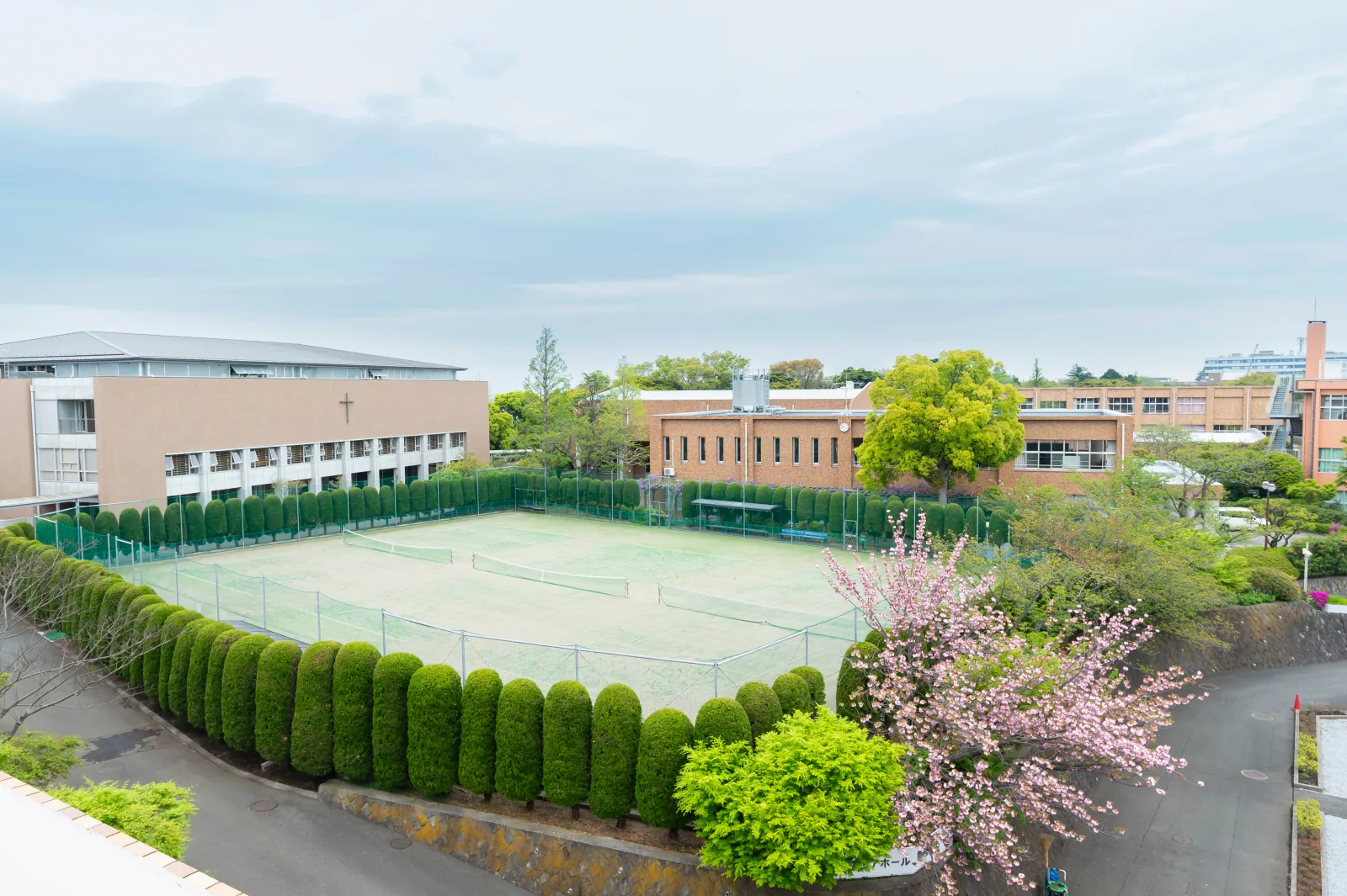 聖園女学院中学校・高等学校のキャンパス全景。緑の生け垣に囲まれたテニスコートの奥に、十字架を掲げた校舎が建っている