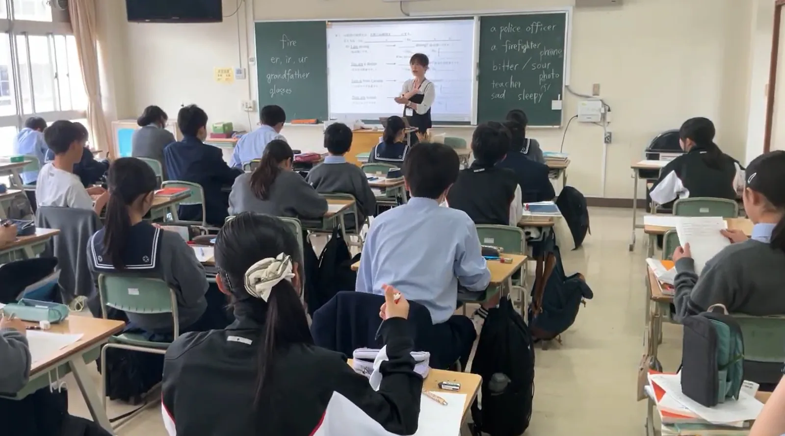 昭和学院秀英中学校・高等学校の英語の授業。黒板と電子黒板の前で先生が説明し、生徒が席でノートをとっている