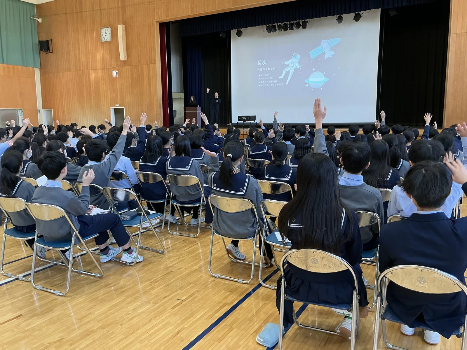 昭和学院秀英中学校・高等学校の講演会。スクリーンの前で話す講師に向かって、いすに座った多くの生徒が手を挙げている
