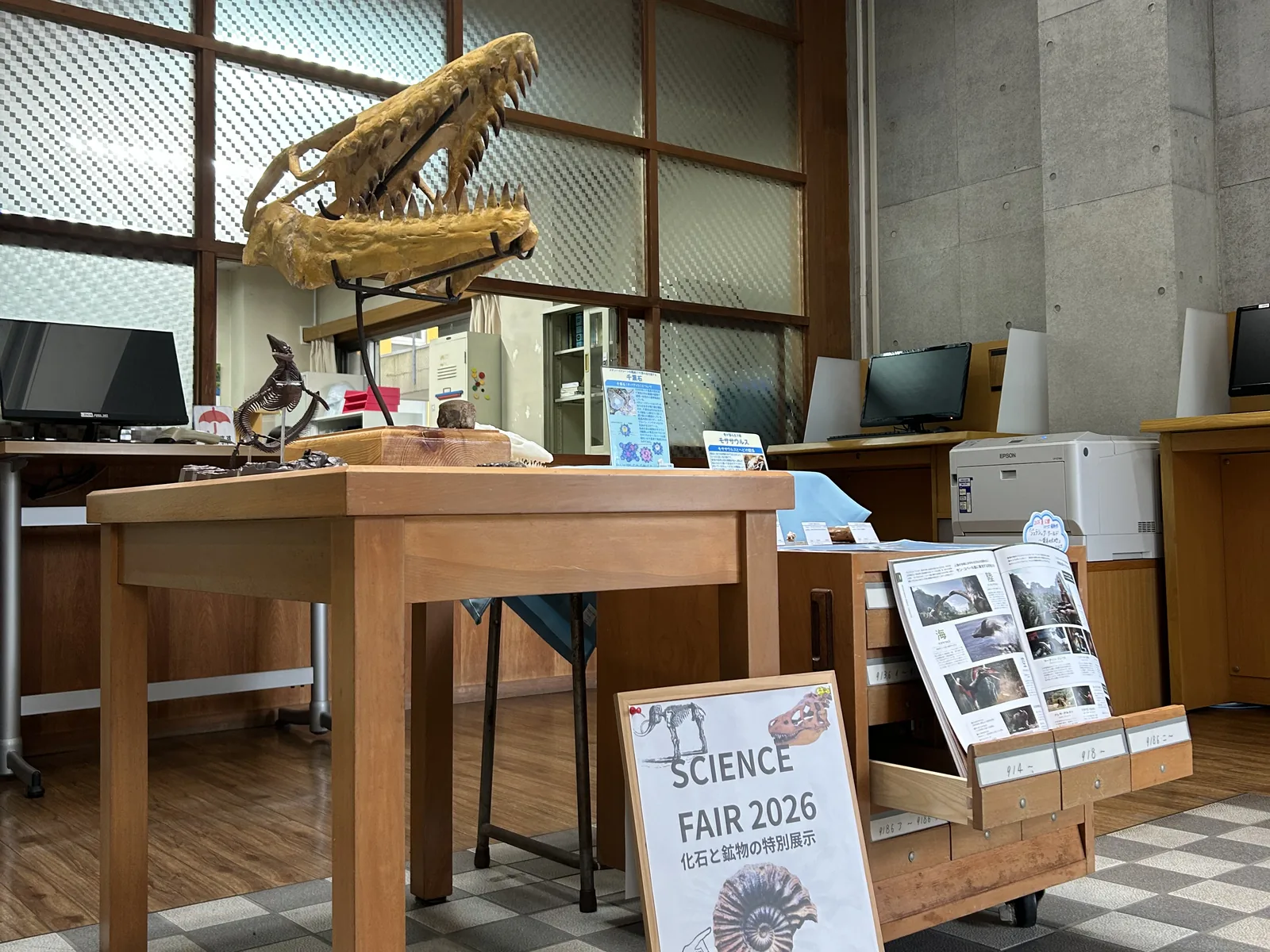 昭和学院秀英中学校・高等学校の図書館。恐竜の頭骨の化石の模型と「SCIENCE FAIR 2026 化石と鉱物の特別展示」のポスターが置かれている