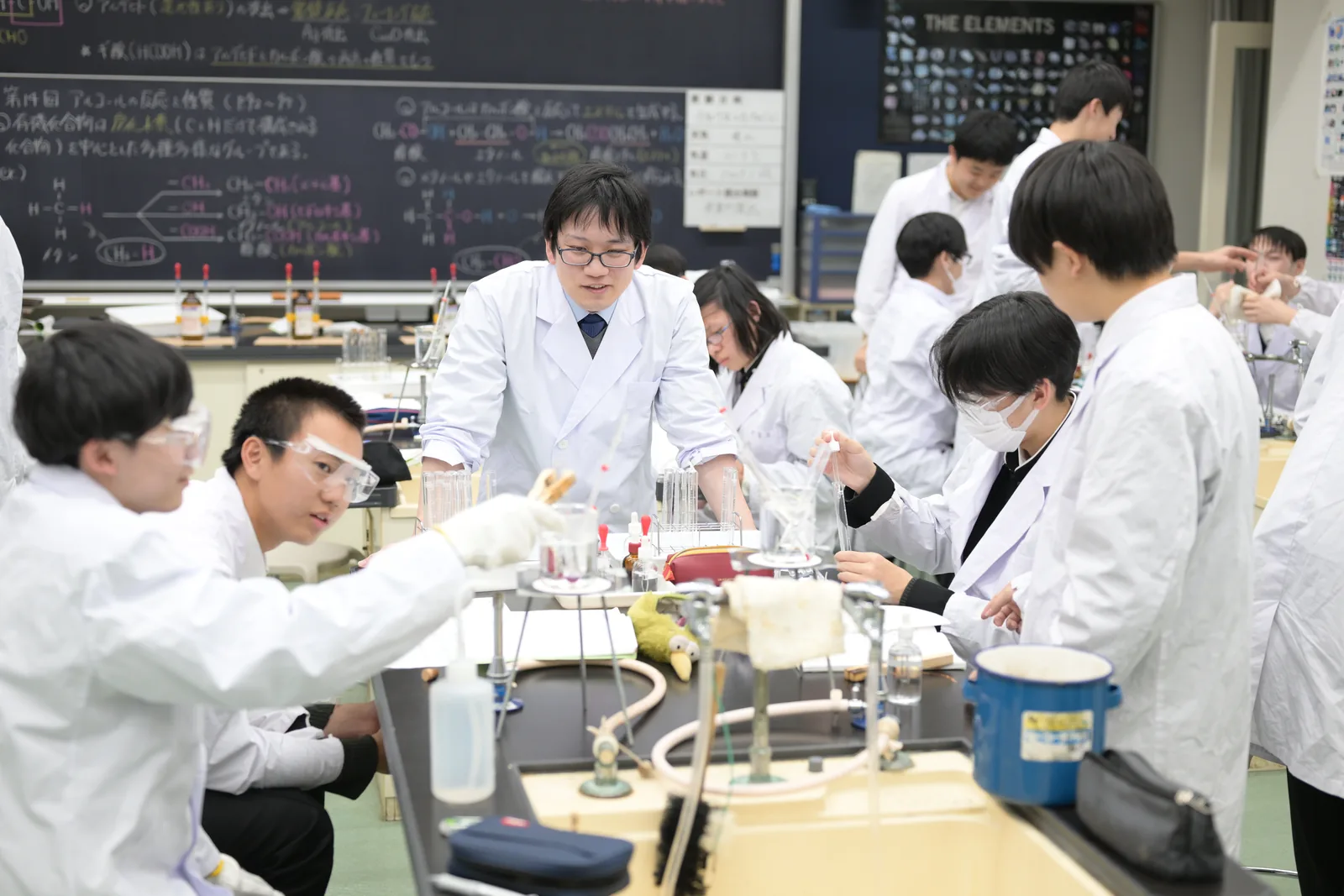 東京都市大学付属中学校・高等学校の理科の実験の授業。白衣と保護眼鏡を着けた生徒が、実験台でガラス器具を使って実験している