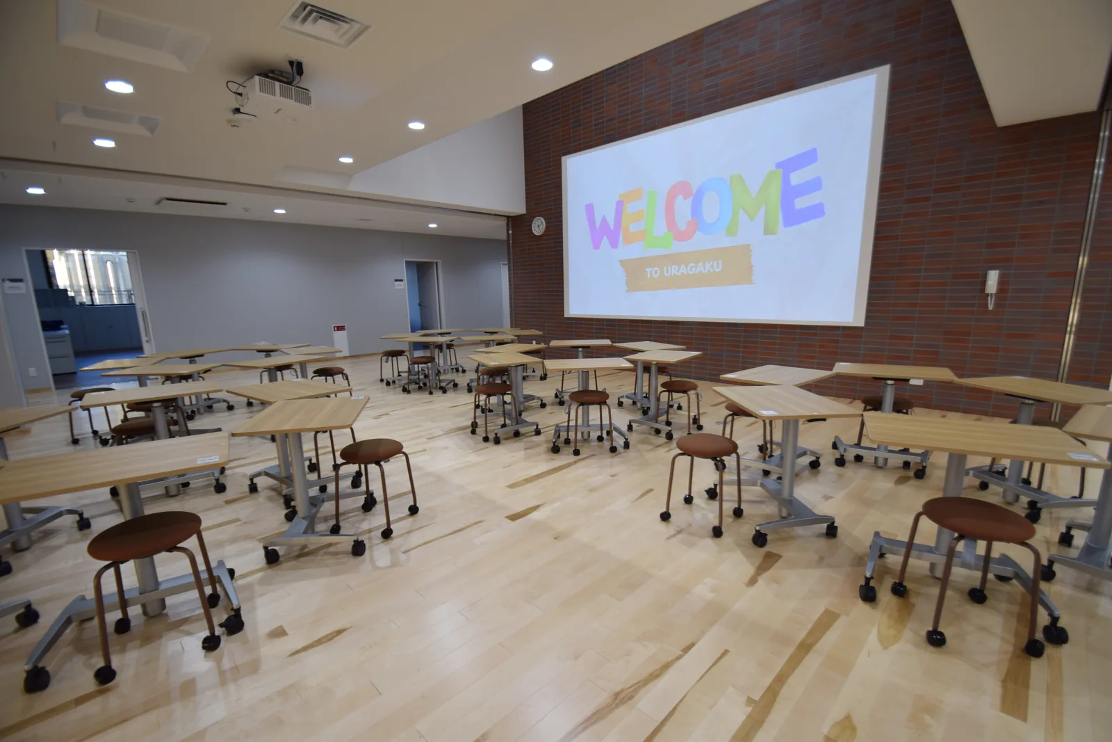 浦和学院中学校のイノベーションコーナー。キャスター付きの机とスツールが並び、正面のスクリーンに映像が映っている